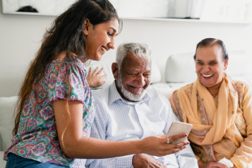 A photo of a young woman sat on a sofa next to an older couple, the young woman shows them something on her phone as they all smile.