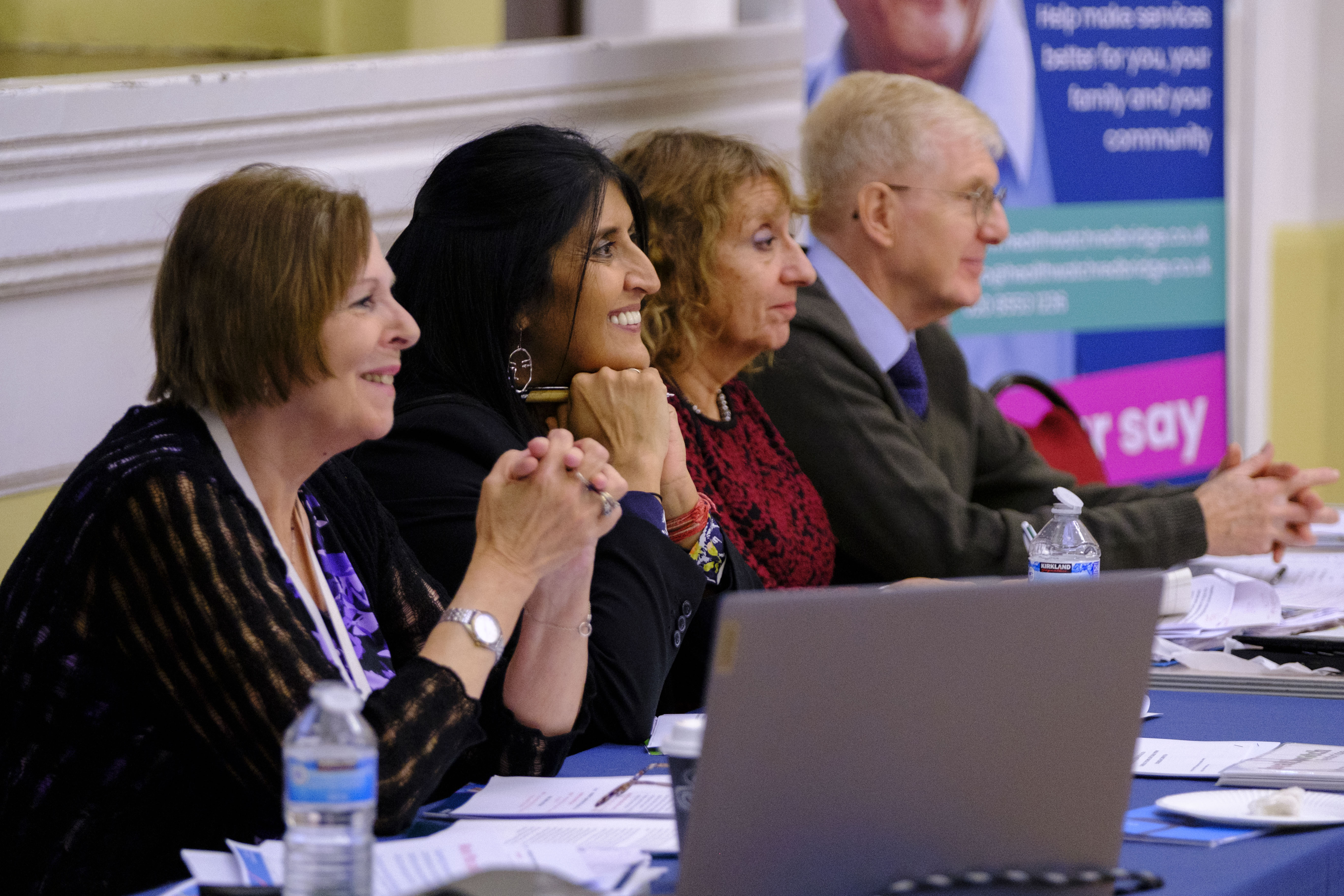 A photograph of four people sitting at a conference table looking out at a seated crowd. 