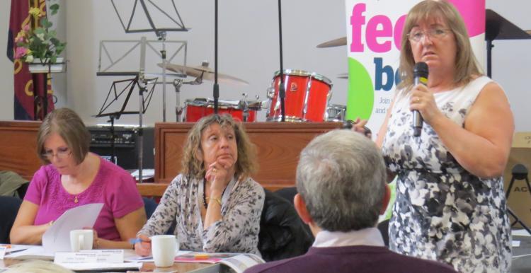 A photo of a woman in a conference space addressing members of a meeting. 