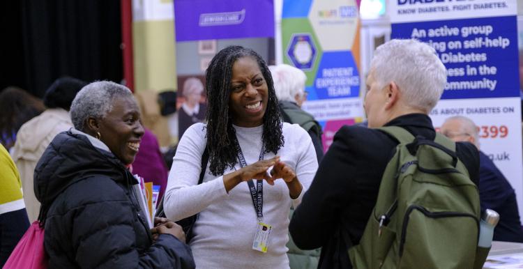 A photo of 3 people stood in a large public hall surrounded by community stalls. A woman in the middle communicates to the group with British Sign Language. 