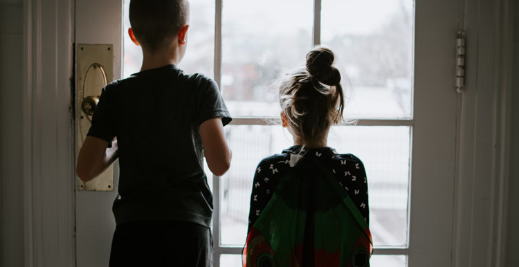 A boy and girl inside a house lookinhg out through the glass door onto the street