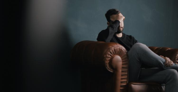 Young man sat on sofa resting head on his hand, looking despondent  