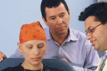 A woman with no hair wearing a head scarf, with her husband standing behind her 