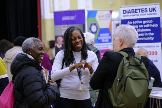 A photo of 3 people stood in a large public hall surrounded by community stalls. A woman in the middle communicates to the group with British Sign Language. 