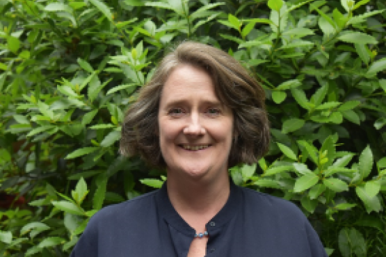 A photo of a smiling woman stood in front of a leafy plant