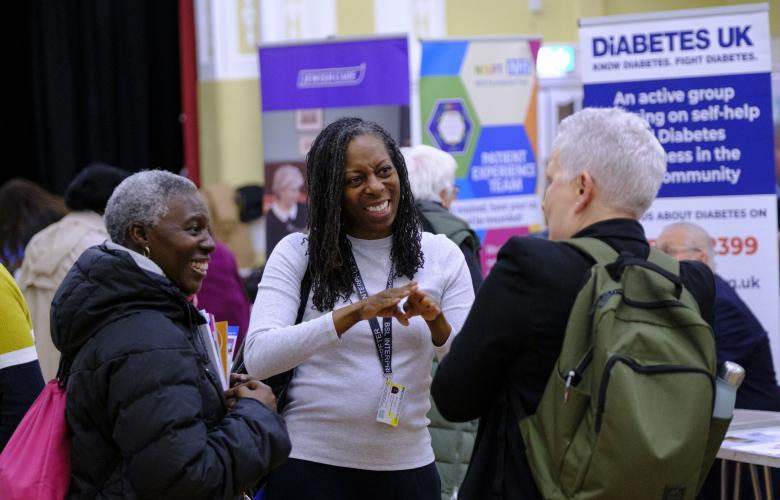 A photo of 3 people stood in a large public hall surrounded by community stalls. A woman in the middle communicates to the group with British Sign Language. 